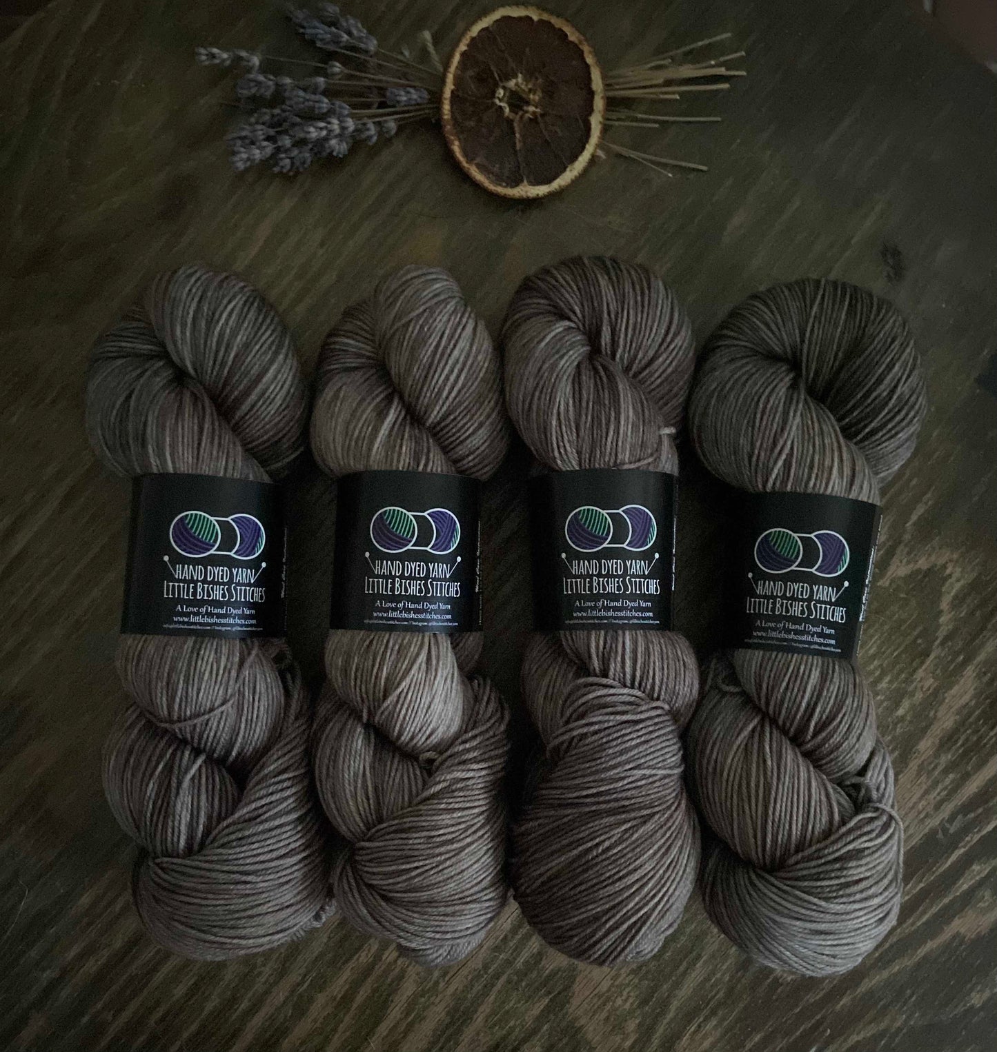 Four skeins of hand-dyed sock yarn in dark colors, arranged side by side on a wooden surface, with a small bundle of dried lavender flowers and a slice of orange to the side.